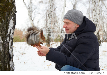 A man sits by a tree in a snowy forest. He reaches out to feed a squirrel some nuts. The squirrel perches on his hand. 134898514