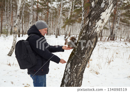 Man feeds squirrel while standing by tree in snowy forest during winter Man feeds squirrel while standing by tree in snowy forest during winter 134898519