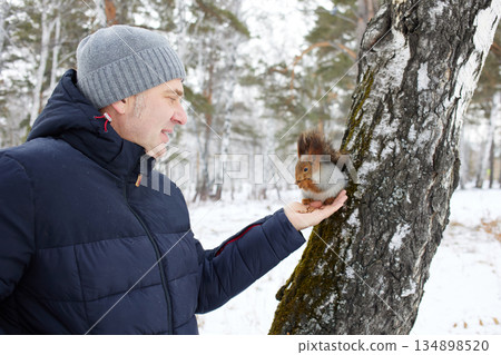 Man feeds squirrel while standing by tree in snowy forest during winter 134898520