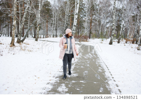 Woman walking on a snowy path in winter with a cup of drink in a forest setting 134898521