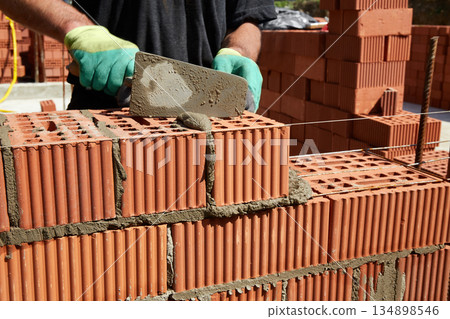 A skilled bricklayer is carefully applying mortar to red bricks, aligning them precisely to build a wall at a construction site A skilled bricklayer is carefully applying mortar to red bricks, aligning them precisely to build a wall at a construction site 134898546