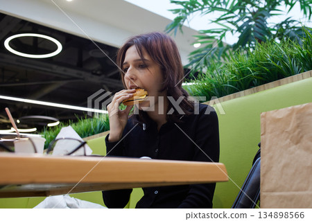A young woman sits at a table in a restaurant and takes a bite of a sandwich. 134898566