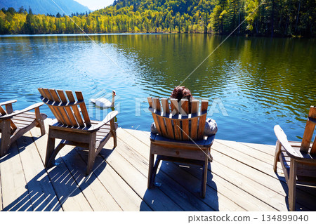 A girl is resting on a wooden sun lounger on the shore of an Emerald Lake on a warm autumn day. A girl is resting on a wooden sun lounger on the shore of an Emerald Lake on a warm autumn day. 134899040