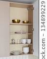 Open kitchen cabinet with brass bowls and white crockery 134899229