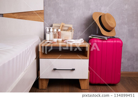 A stylish bedside table with personal hygiene items and a bright pink suitcase with a straw hat A stylish bedside table with personal hygiene items and a bright pink suitcase with a straw hat 134899451