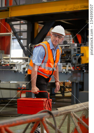 Industrial technician wearing vest safety crouching beside factory machinery with red toolbox, preparing for maintenance. Concept of workplace safety, repair and factory equipment servicing. 134899507