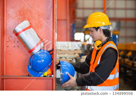 Industrial worker in safety vest and helmet placing blue hard hat on storage shelf inside factory. Concept of workplace organization, safety compliance and proper PPE management in industry. Industrial worker in safety vest and helmet placing blue hard hat on storage shelf inside factory. Concept of workplace organization, safety compliance and proper PPE management in industry. 134899508