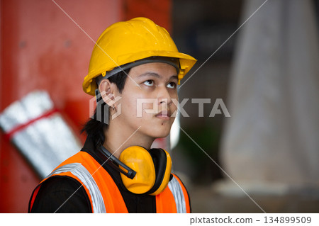 Close-up of young factory worker wearing safety helmet, ear protection, and reflective vest, looking upward with focus. Concept of industrial safety, awareness, and concentration in workplace. 134899509
