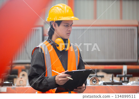 Young Asian factory worker wearing safety vest, helmet, and earmuffs holding clipboard checking for inspection in industrial environment. Concept of safety, engineering and manufacturing. Young Asian factory worker wearing safety vest, helmet, and earmuffs holding clipboard checking for inspection in industrial environment. Concept of safety, engineering and manufacturing. 134899511