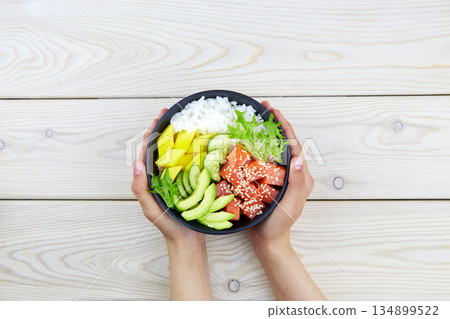 Poke bowl of salmon, rice, avocado, cucumber, mango, herbs in a woman's hands. Poke bowl of salmon, rice, avocado, cucumber, mango, herbs in a woman's hands. 134899522