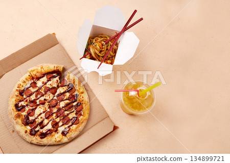 A tempting round pizza topped with cheese and slices of pepperoni sits next to a takeout container filled with colorful noodles. 134899721