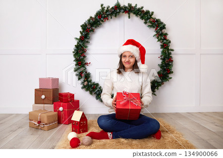 A happy girl in a white knitted sweater and a Santa Claus hat opens a Christmas gift A happy girl in a white knitted sweater and a Santa Claus hat opens a Christmas gift 134900066