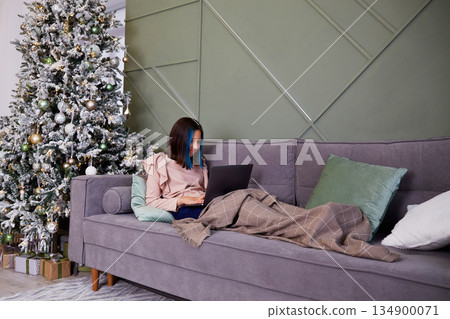 A pretty girl is working on a laptop, covered with a blanket. Christmas tree with toys and gifts. A pretty girl is working on a laptop, covered with a blanket. Christmas tree with toys and gifts. 134900071