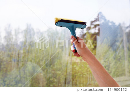 A young women is cleaning a large window with a squeegee, revealing a scenic outdoor view A young women is cleaning a large window with a squeegee, revealing a scenic outdoor view 134900152