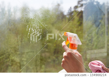 A hand holds a spray bottle, applying cleaner to a window.Window cleaning. A hand holds a spray bottle, applying cleaner to a window.Window cleaning. 134900214