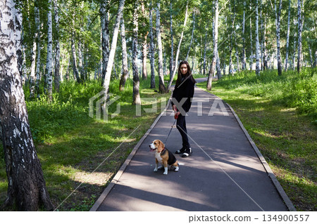 A girl with a beagle dog walks along a shady path in a park surrounded by green trees during the daytime A girl with a beagle dog walks along a shady path in a park surrounded by green trees during the daytime 134900557