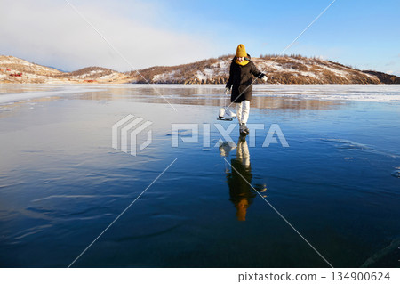 A happy girl looks at her reflection on the ice of the frozen Lake Baikal. He holds skates in his hands. A happy girl looks at her reflection on the ice of the frozen Lake Baikal. He holds skates in his hands. 134900624