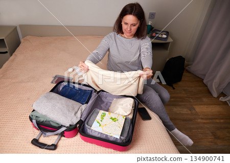 A woman is sitting on a bed, packing a suitcase for an upcoming trip, folding clothes and travel supplies 134900741