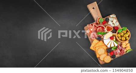 Antipasto platter with prosciutto, cheeses, olives, figs and crackers on wooden board over black background with copy space Antipasto platter with prosciutto, cheeses, olives, figs and crackers on wooden board over black background with copy space 134904850