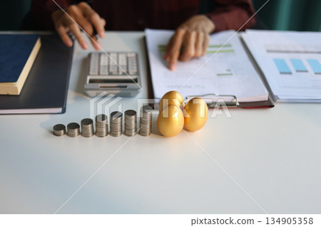 Rising stack of coins and golden eggs on the table. Businessman calculating dividend yield on the background for wealth management and investment concept. 134905358