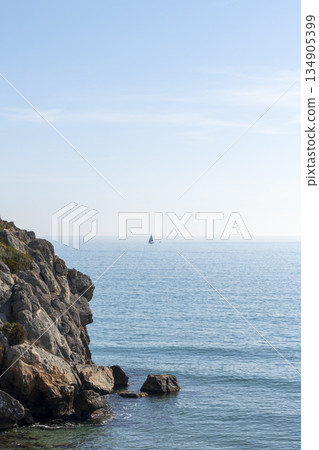rocky coastline with a sailboat sailing with a calm sea and the horizon in the background 134905399