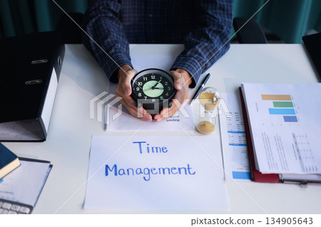 Time management concept. Businessman working on office desk with stack of documents, running sand clock and clock on the table. Time management concept. Businessman working on office desk with stack of documents, running sand clock and clock on the table. 134905643