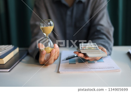 Time and money concept. Businessman holding cash and sand clock imply growth of income, saving or return investment through time. Time and money concept. Businessman holding cash and sand clock imply growth of income, saving or return investment through time. 134905699