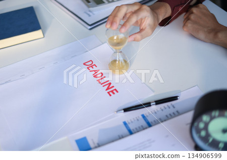 Male employee sitting on office desk holding running sand clock and Deadline paper on the table. Time management concept. Male employee sitting on office desk holding running sand clock and Deadline paper on the table. Time management concept. 134906599