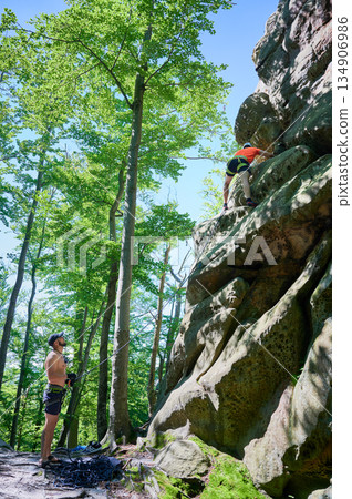 Male rock climber ascending rugged limestone cliff with harness and rope for safety. Sportsman climbing on vertical large boulder at Dobvush Rocks in Carpathian mountains, Ukraine. Male rock climber ascending rugged limestone cliff with harness and rope for safety. Sportsman climbing on vertical large boulder at Dobvush Rocks in Carpathian mountains, Ukraine. 134906986