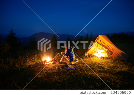 Woman hiker sits by campfire near her illuminated tent in soft glow of twilight, surrounded by serene mountain scenery. Clear night sky above adds peaceful ambiance to quiet wilderness setting. 134906990