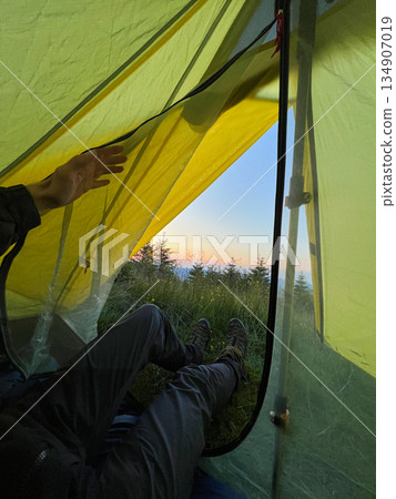 Camper peeks out from yellow tent at dawn, revealing serene view of grassy terrain and distant trees. Dewdrops glisten on tent fabric, with hiking boots placed at tent's entrance. Camper peeks out from yellow tent at dawn, revealing serene view of grassy terrain and distant trees. Dewdrops glisten on tent fabric, with hiking boots placed at tent's entrance. 134907019