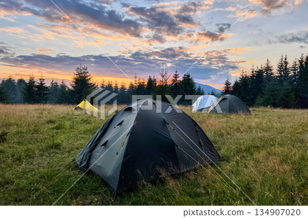 Few tourist tents in grassy field in evening, surrounded by trees and hills under stunning sunset sky. Colorful clouds enhance beauty of scene, with hues of orange and purple illuminating horizon. Few tourist tents in grassy field in evening, surrounded by trees and hills under stunning sunset sky. Colorful clouds enhance beauty of scene, with hues of orange and purple illuminating horizon. 134907020