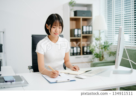 A focused office woman writing notes and working on paperwork at her desk, representing productivity, organization, business tasks, 134911511
