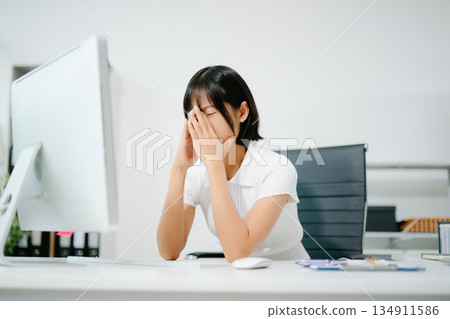 A stressed office woman holding her head with fatigue and headache, showing workplace pressure, mental strain, and emotional exhaustion from overworking. 134911586