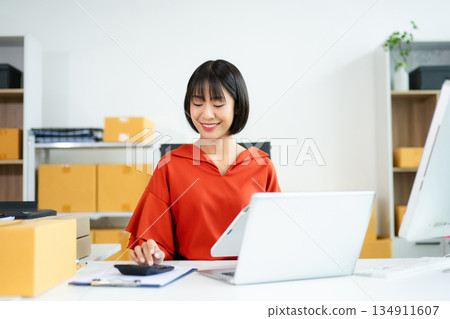 Young Asian woman using calculator and laptop in modern office with boxes. Ideal for themes like e-commerce, small business, shipping, startup 134911607