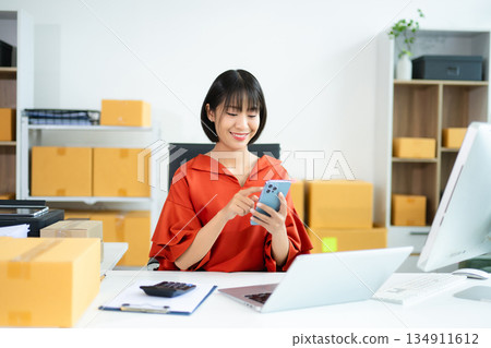 Young Asian woman using tablet in e-commerce office with boxes, laptop, and smile. Perfect for business, shipping, startup, and online seller concepts. Young Asian woman using tablet in e-commerce office with boxes, laptop, and smile. Perfect for business, shipping, startup, and online seller concepts. 134911612