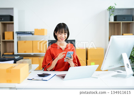 Entrepreneurial Asian woman prepares parcels and checks online orders on tablet and laptop for delivery online shopping and small business success in office 134911615