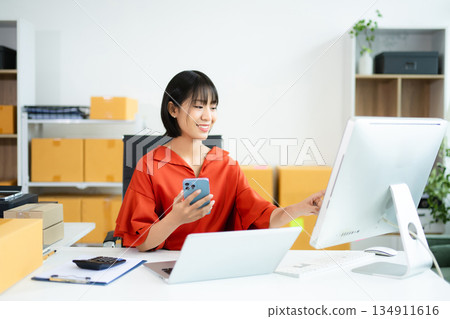 Young Asian woman using tablet in e-commerce office with boxes, laptop, and smile. Perfect for business, shipping, startup, and online seller concepts. Young Asian woman using tablet in e-commerce office with boxes, laptop, and smile. Perfect for business, shipping, startup, and online seller concepts. 134911616