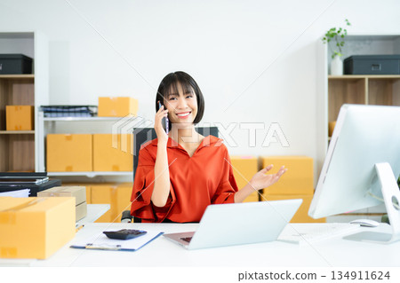 Smiling Asian entrepreneur talks on phone and holds package in modern office. Smiling Asian entrepreneur talks on phone and holds package in modern office. 134911624