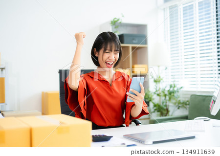 Smiling Asian woman raises fist in joy while holding smartphone. Perfect for business success, ecommerce wins, startup celebration, and real people themes. Smiling Asian woman raises fist in joy while holding smartphone. Perfect for business success, ecommerce wins, startup celebration, and real people themes. 134911639