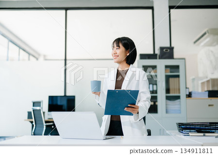 Smiling Asian woman in white blazer holding coffee cup and clipboard in bright office. Confident, stylish businesswoman balancing work and lifestyle. Smiling Asian woman in white blazer holding coffee cup and clipboard in bright office. Confident, stylish businesswoman balancing work and lifestyle. 134911811