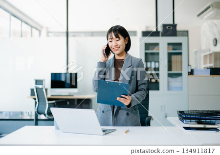Asian Business woman Talking on the phone and using a laptop with a smile while sitting at office 134911817