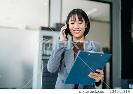 Asian Business woman Talking on the phone and using a laptop with a smile while sitting at office Asian Business woman Talking on the phone and using a laptop with a smile while sitting at office 134911819