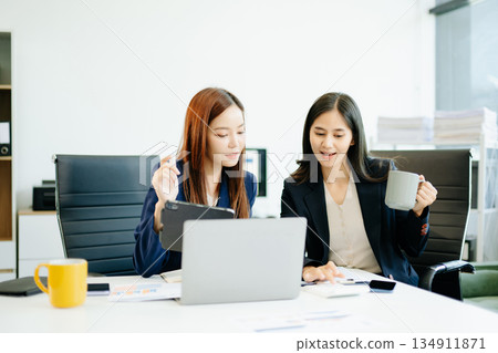 Two Asian businesswomen in suits analyze reports together at a modern office desk. A scene of teamwork, leadership 134911871