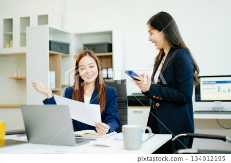 Two Asian businesswomen in suits analyze reports together at a modern office desk. A scene of teamwork, leadership Two Asian businesswomen in suits analyze reports together at a modern office desk. A scene of teamwork, leadership 134912195
