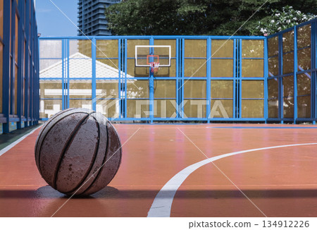 On the orange cement floor of the outdoor court, an old basketball rests in the foreground, with a basketball hoop, fiberglass backboard, and blue steel fence visible behind it. On the orange cement floor of the outdoor court, an old basketball rests in the foreground, with a basketball hoop, fiberglass backboard, and blue steel fence visible behind it. 134912226