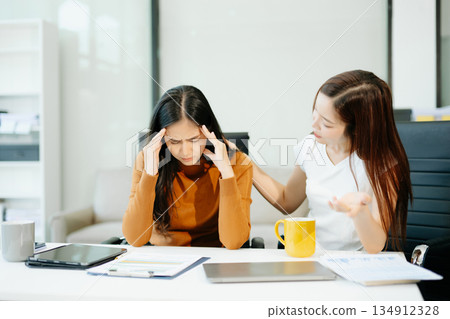 Asian woman comforts her distressed coworker in office. Concept of workplace stress, burnout, empathy, and emotional support in professional environments. Asian woman comforts her distressed coworker in office. Concept of workplace stress, burnout, empathy, and emotional support in professional environments. 134912328