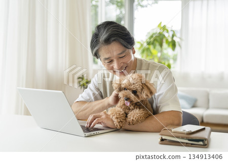 一位五十多歲的男子一邊用筆記型電腦工作,一邊抱著他的寵物狗 一位五十多歲的男子一邊用筆記型電腦工作,一邊抱著他的寵物狗 134913406