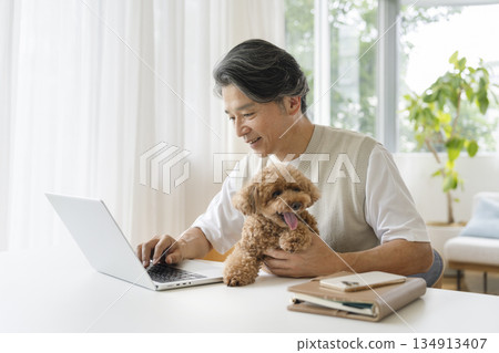 A man in his 50s working on a laptop while holding his pet dog 134913407