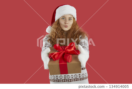 Curly redhead girl in Santa hat and sweater is giving a present and kiss on red background. Curly redhead girl in Santa hat and sweater is giving a present and kiss on red background. 134914005